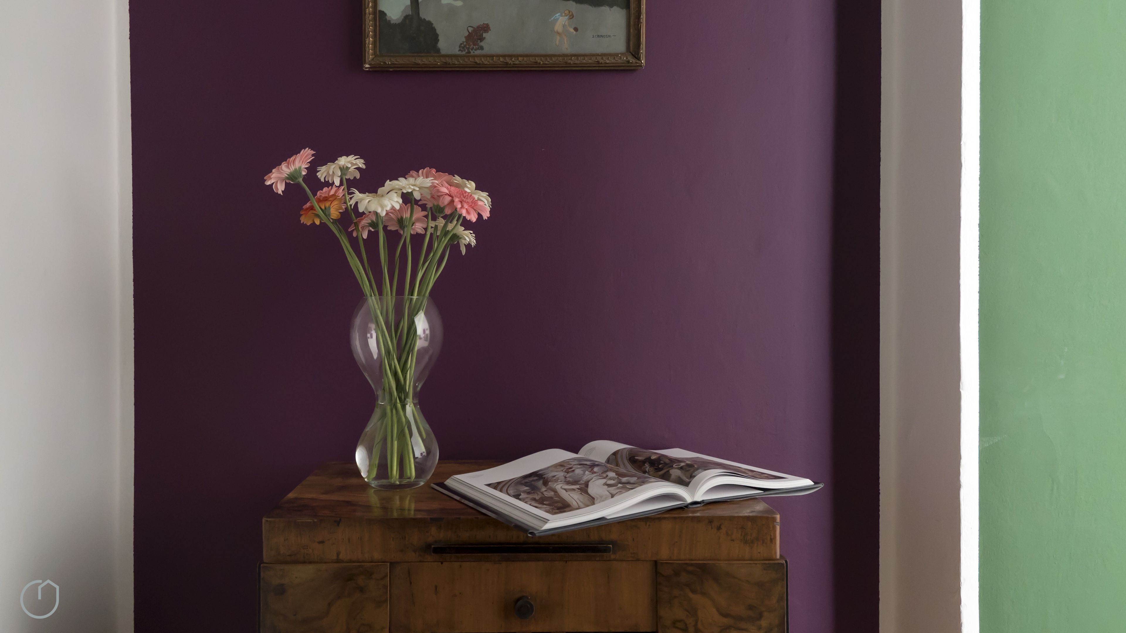 A wooden side table with a clear vase containing flowers, an open magazine, and a framed picture on a purple wall.