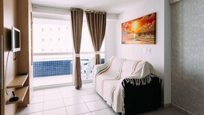 A hotel room with a large window, a TV mounted on the wall, a painting, and a sofa with a throw blanket. - Beach Class - MeuLugarCeará (Fortaleza)