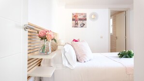 A neatly arranged bedroom with a bed, a bedside table with a vase of flowers, a wall-mounted shelf, and a painting.