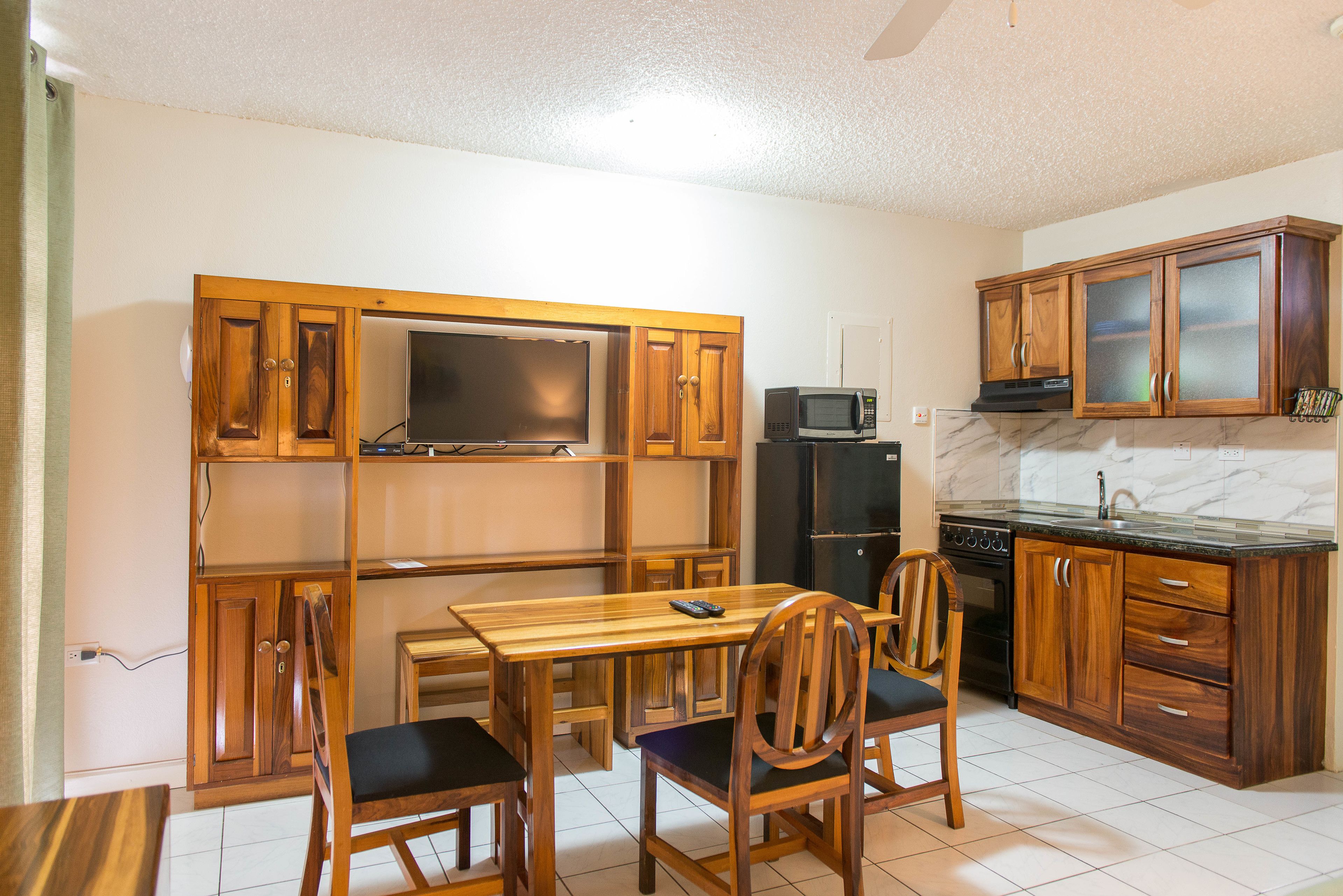 A compact kitchen with wooden cabinets, a built-in microwave, a refrigerator, and a dining table with chairs.