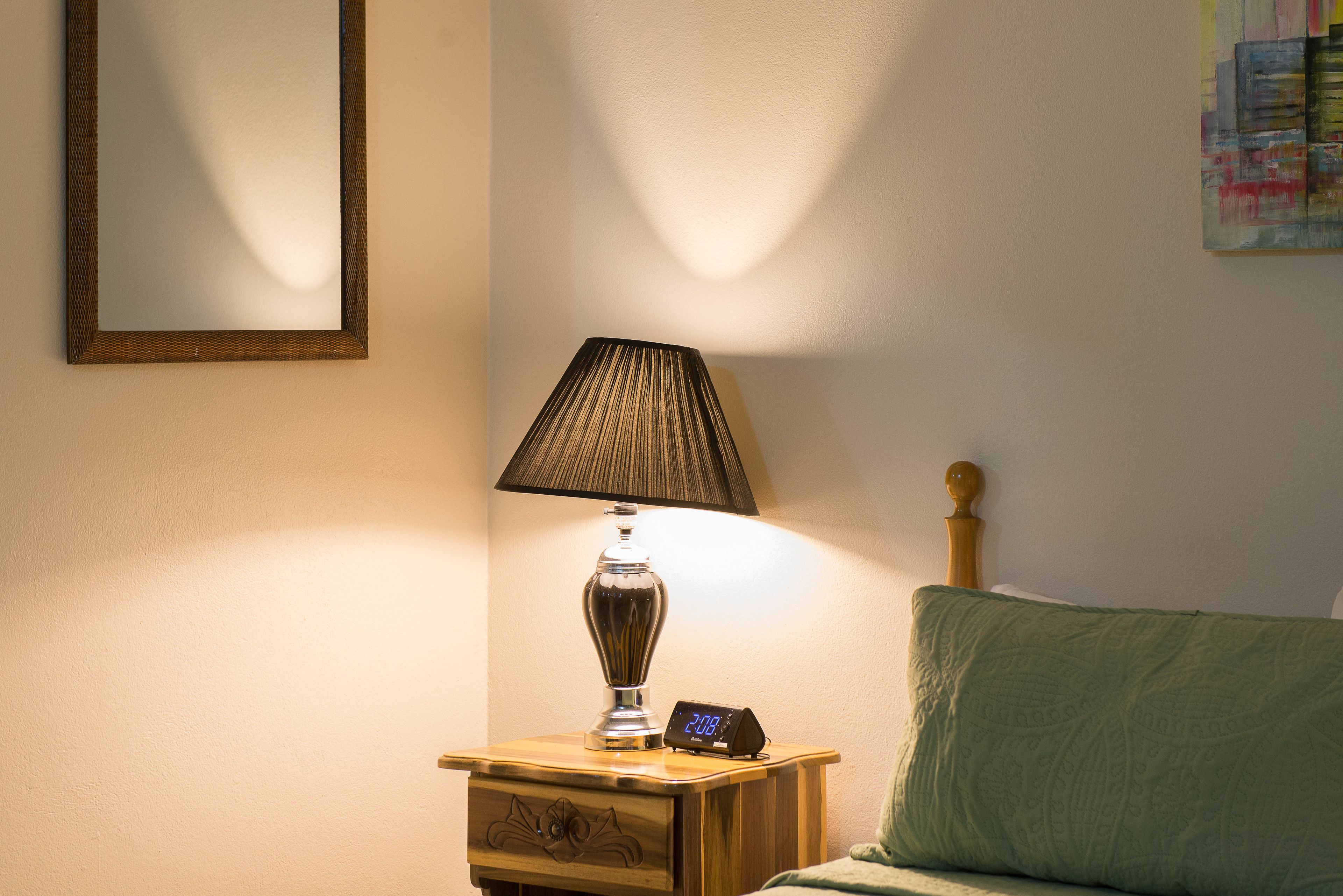 A bedside table with a lamp, a digital clock, and a mirror on the wall.