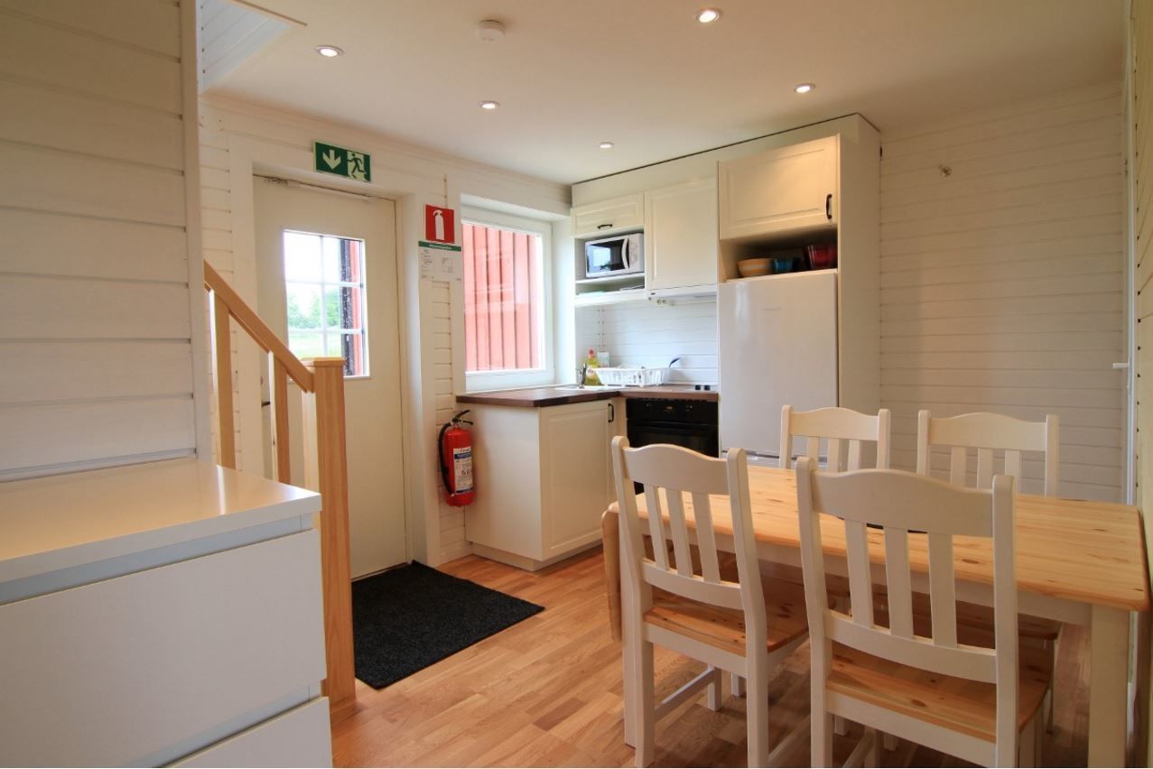 A compact kitchen with white cabinetry, a microwave, and a refrigerator. There is a wooden dining table with chairs and a black doormat on the floor.