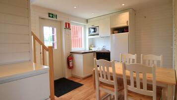 A compact kitchen with white cabinetry, a microwave, and a refrigerator. There is a wooden dining table with chairs and a black doormat on the floor.