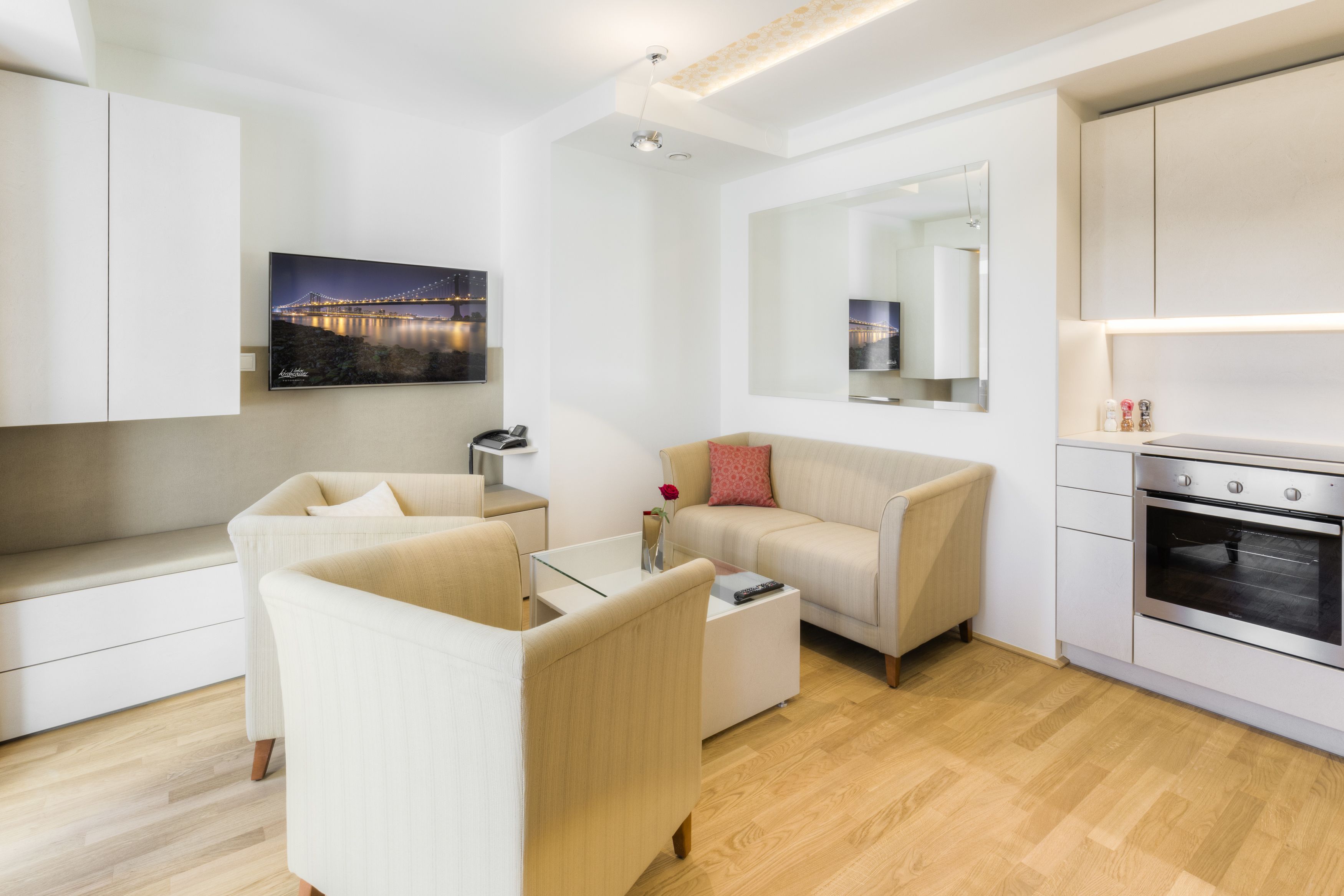 A modern living room with a sofa, a coffee table, and a TV mounted on the wall. There is a kitchen area with an oven and a countertop.