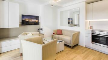 A modern living room with a sofa, a coffee table, and a TV mounted on the wall. There is a kitchen area with an oven and a countertop.