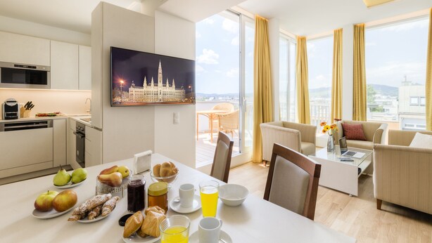 Et moderne hotelværelse med spiseplads, en opholdsstue med sofa og sofabord, et fladskærms-tv og en balkon med udsigt over byen.