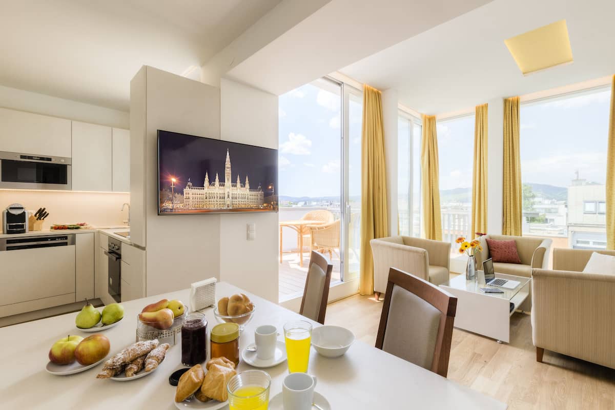 A modern hotel room with a dining area, a living room with a sofa and a coffee table, a flat-screen TV, and a balcony with a city view.