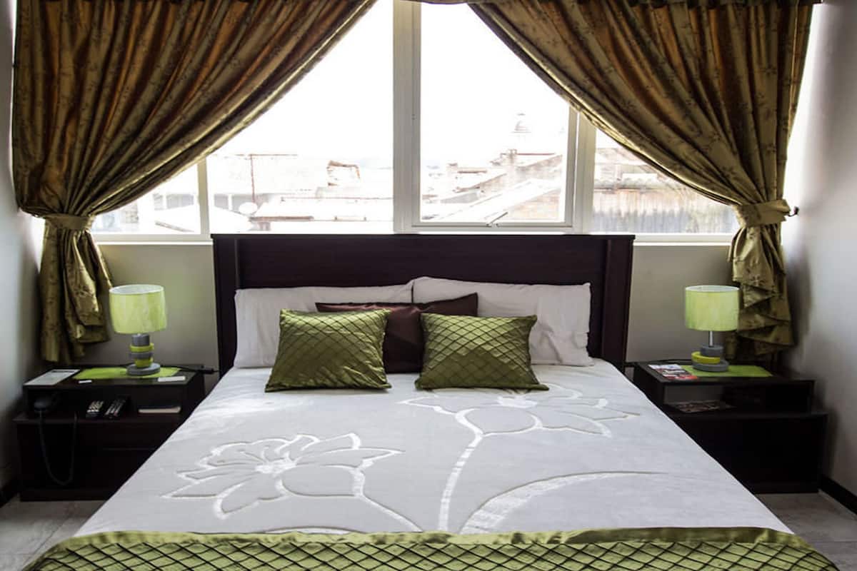 A bedroom with a bed, two nightstands, a window with curtains, and a view of buildings outside.