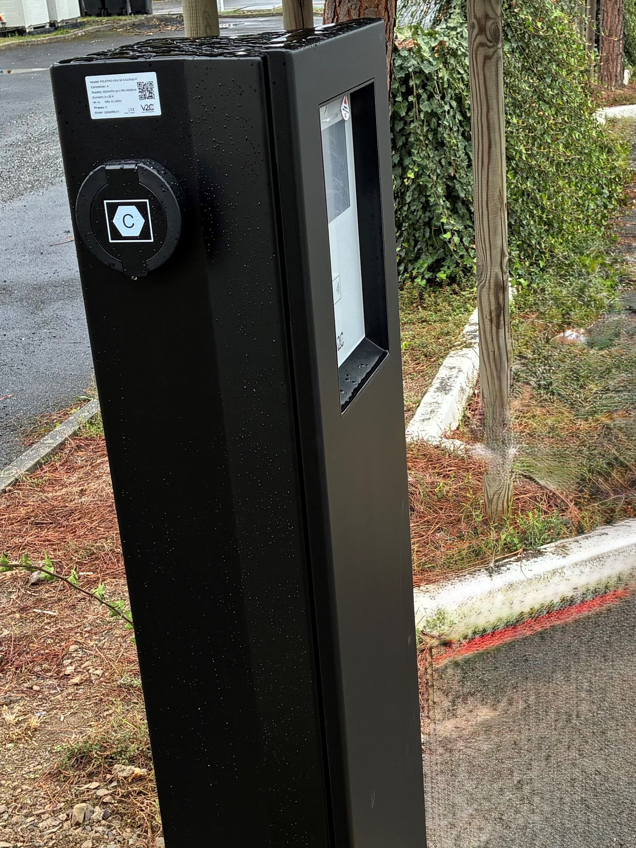 Musta sähkökotelo, jossa on QR-koodi ja etiketti. Se sijaitsee ulkona puun ja jalkakäytävän läheisyydessä.