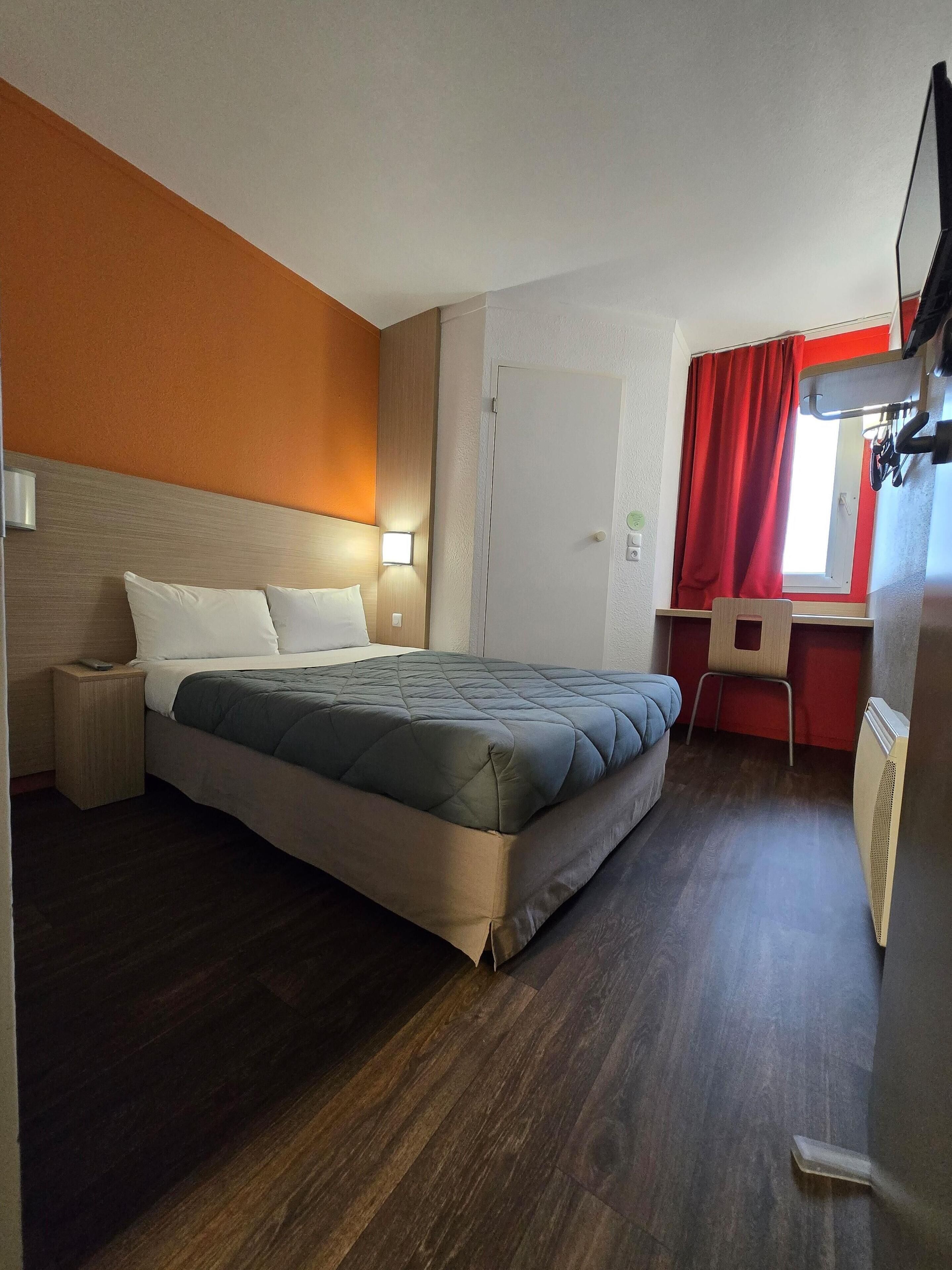 Quarto de hotel com cama, mesa de cabeceira, luminária, escrivaninha, cadeira e janela com cortinas vermelhas.