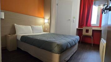 Quarto de hotel com cama, mesa de cabeceira, luminária, escrivaninha, cadeira e janela com cortinas vermelhas.