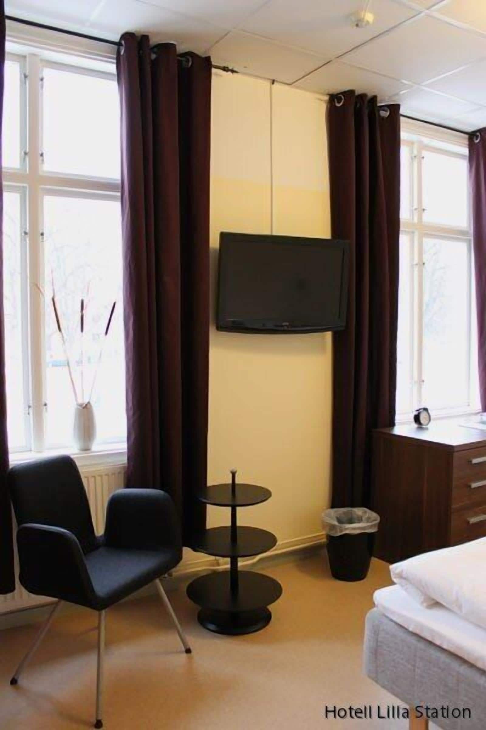 A hotel room with a bed, a TV mounted on the wall, a small round table, a chair, and a window with curtains.
