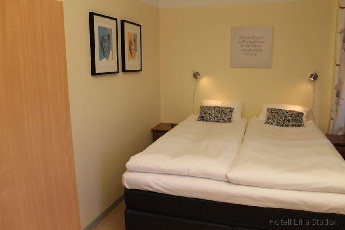 A double bed with white bedding and two decorative pillows, a bedside table, and a wall-mounted light fixture.