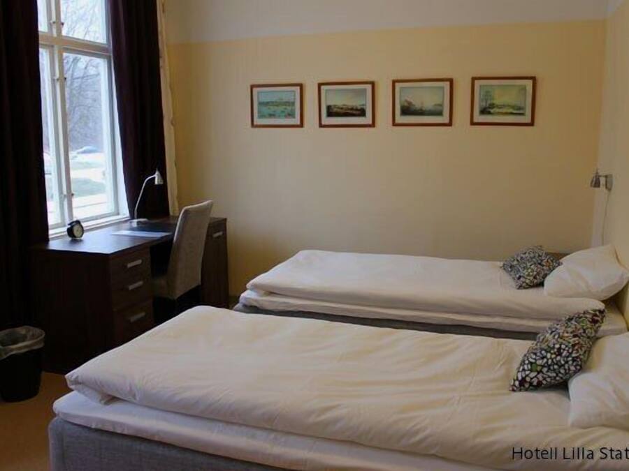 A hotel room with two beds, a desk, and a window. There are four framed pictures on the wall.