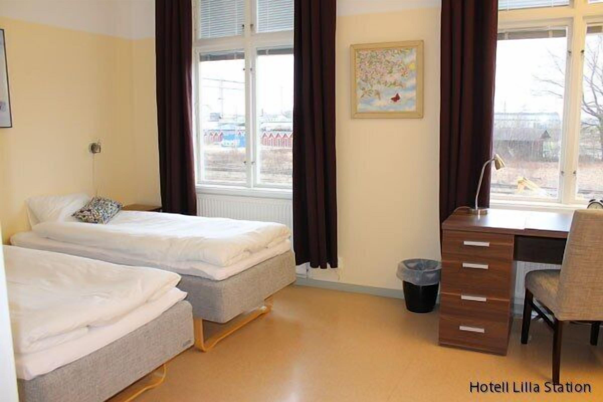 A hotel room with two single beds, a desk, a chair, a radiator, a window with curtains, and a trash bin.