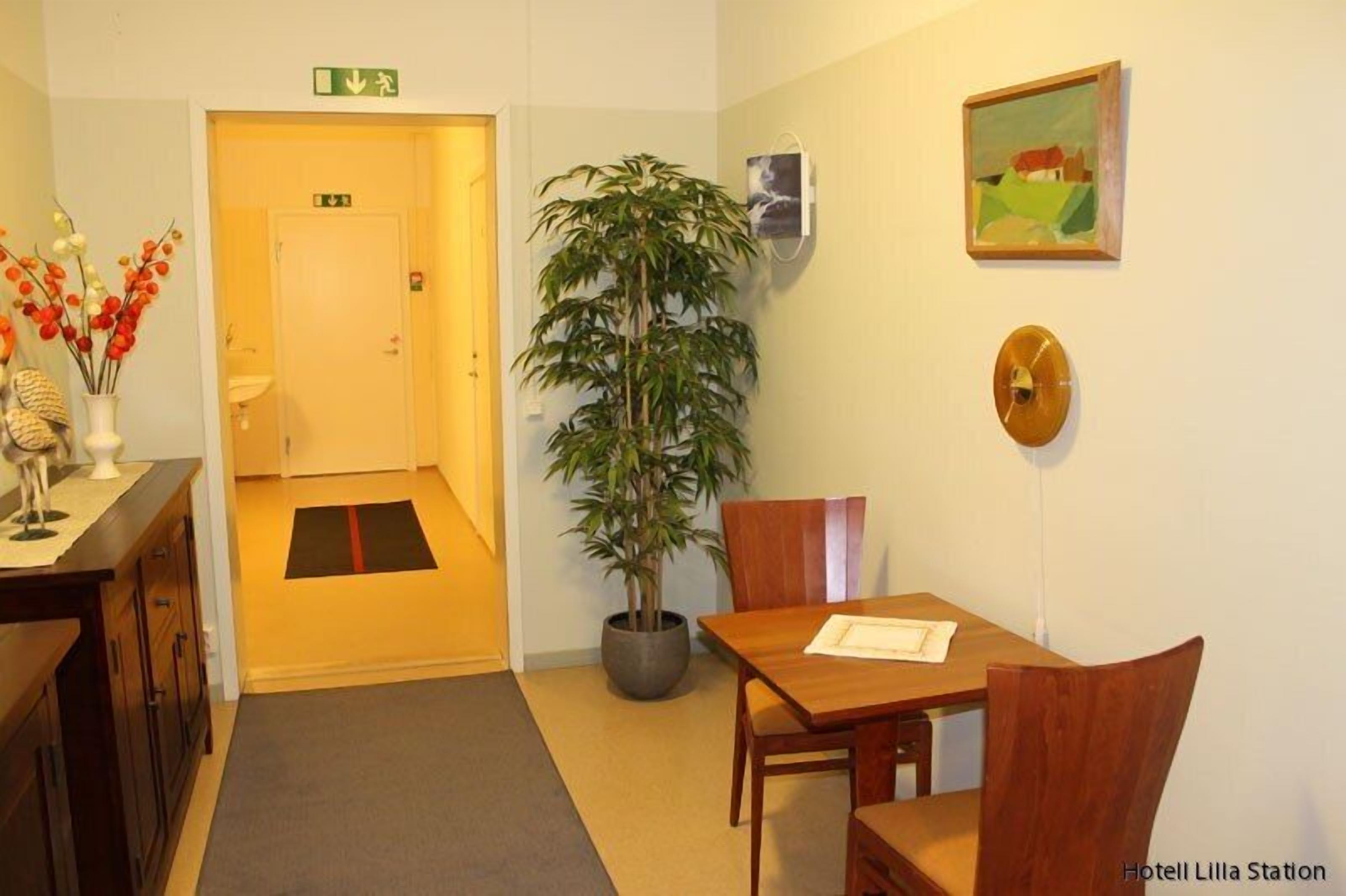 A hotel room with a wooden dining table, chairs, a potted plant, a painting, and a clock on the wall.