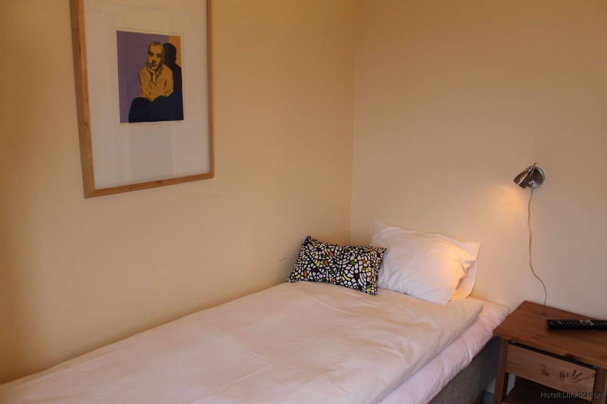 A single bed with white bedding, a framed picture on the wall, and a bedside table with a lamp and remote control.