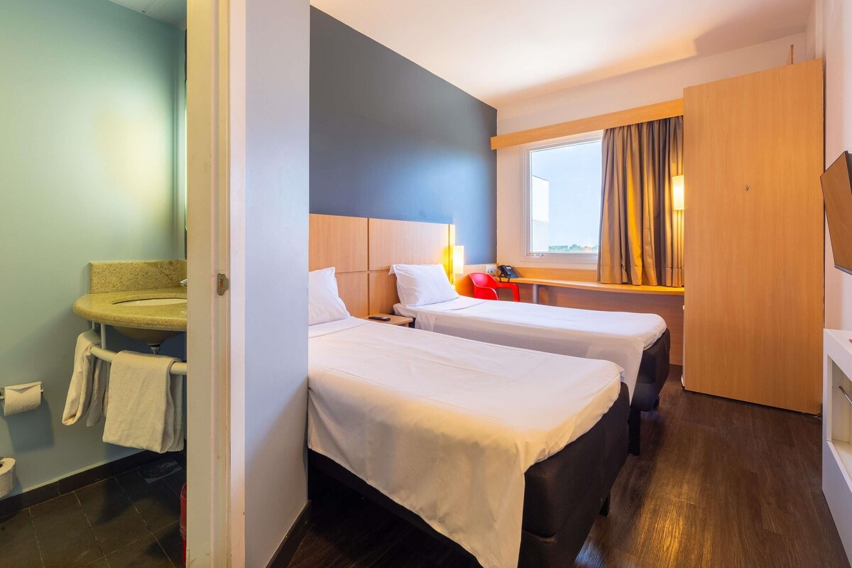 Hotelkamer met twee bedden, een bureau, een raam met gordijnen, en een badkamer met wastafel en handdoekrek.