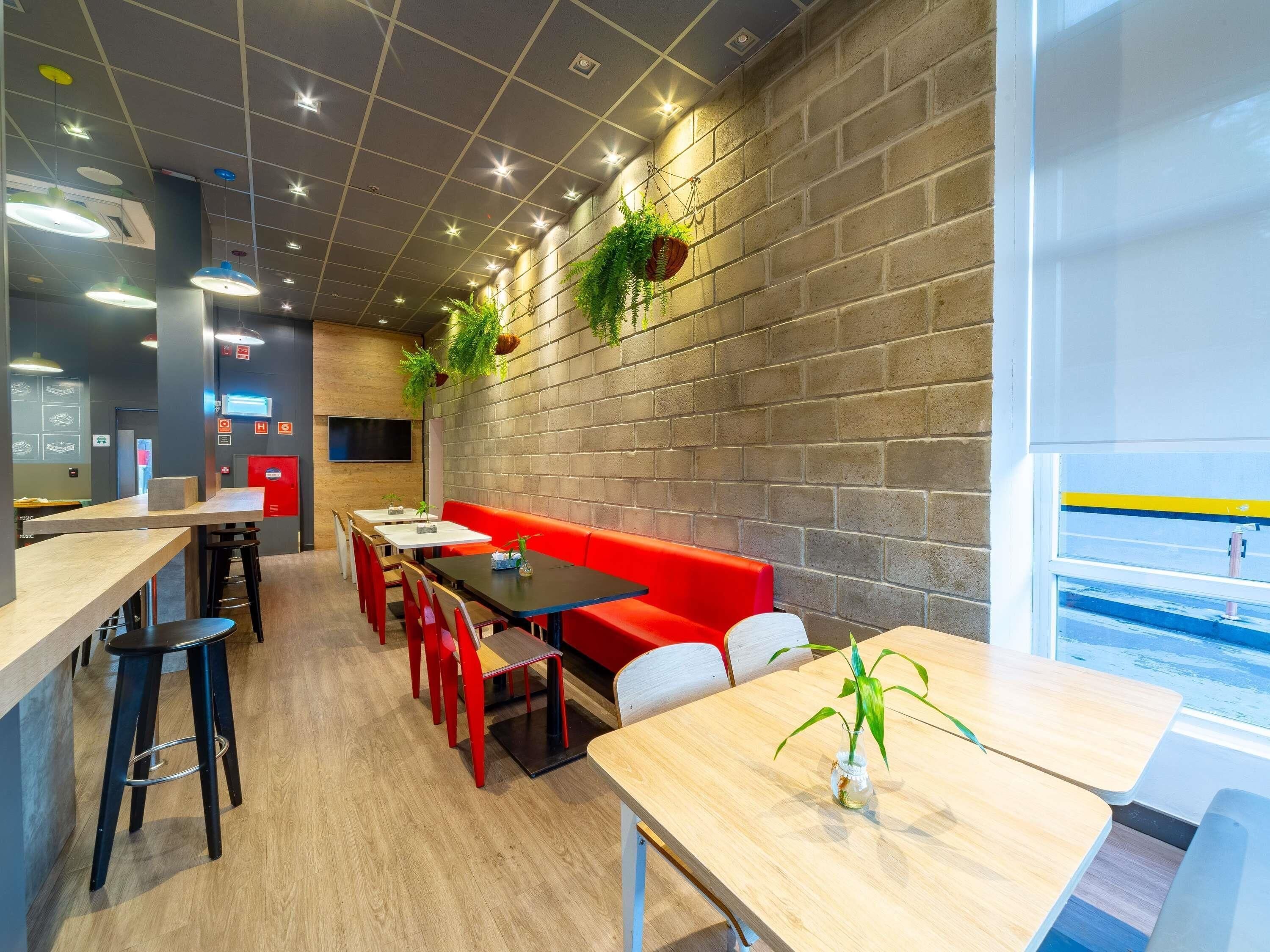 A modern restaurant with a brick wall, wooden tables, red seating, and hanging plants.