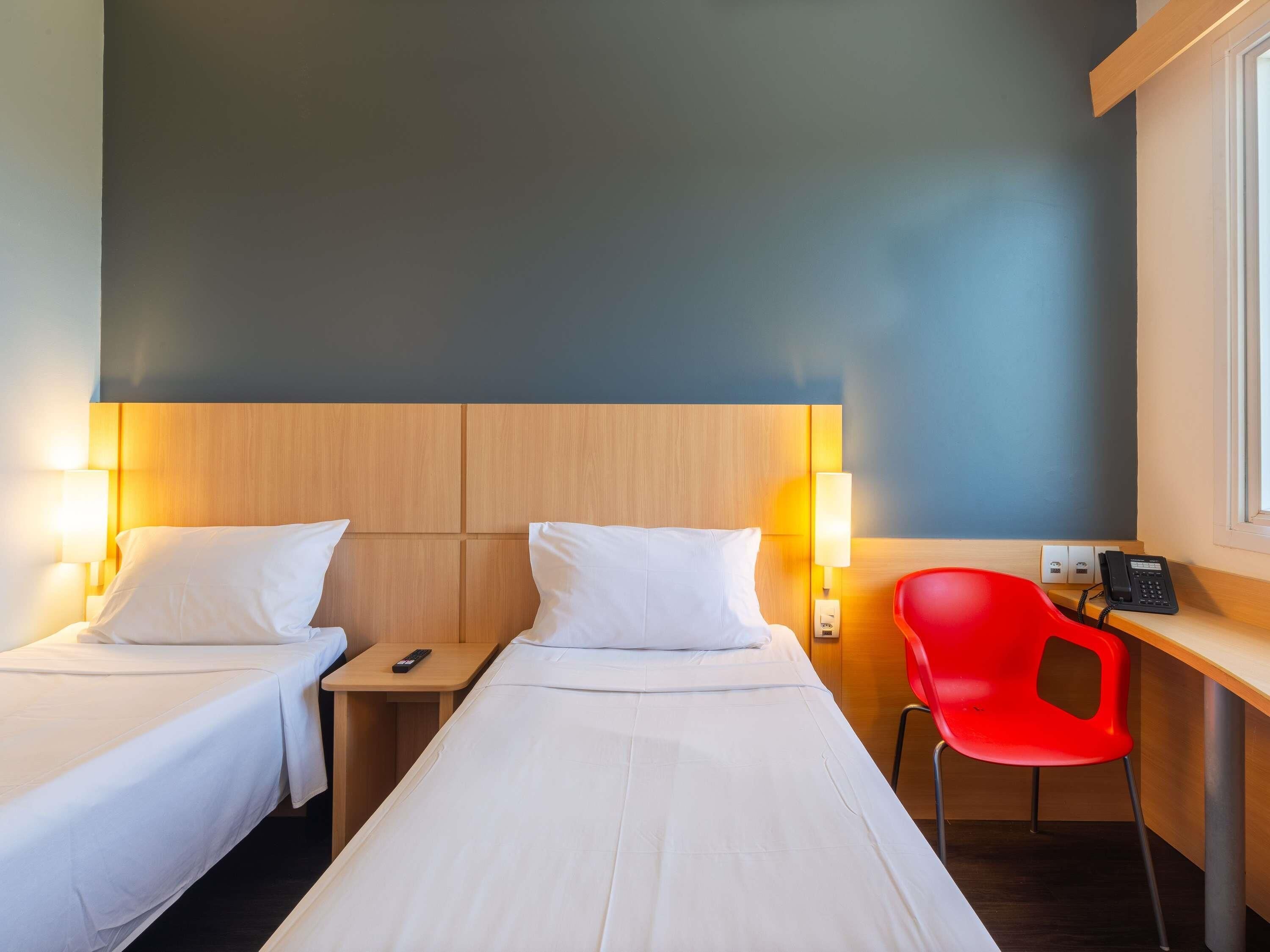 A hotel room with two beds, bedside lamps, a desk with a phone, and a red chair.