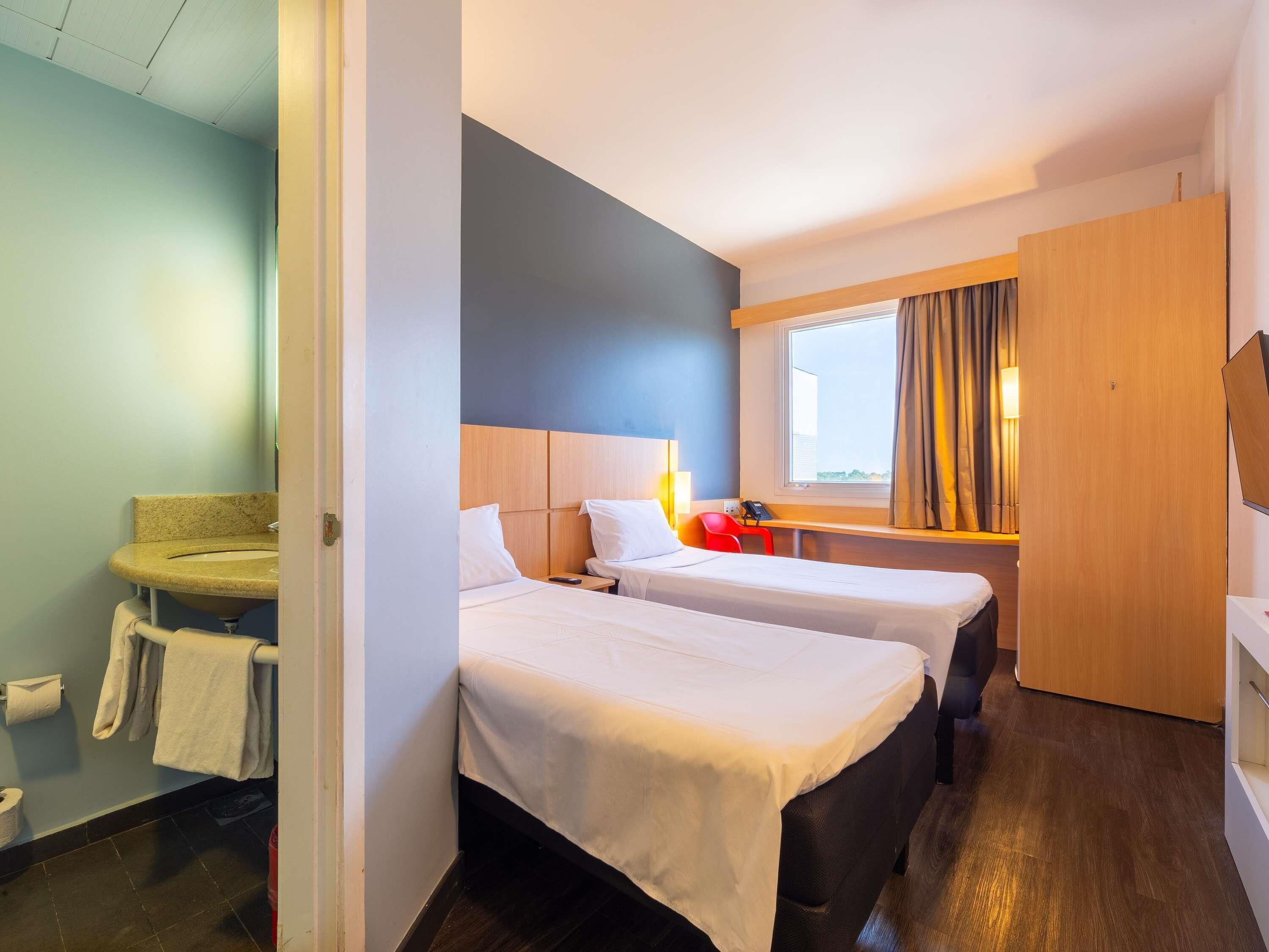 Une chambre d’hôtel avec deux lits, un bureau, une fenêtre avec des rideaux et une salle de bain équipée d’un lavabo et d’un porte-serviettes.