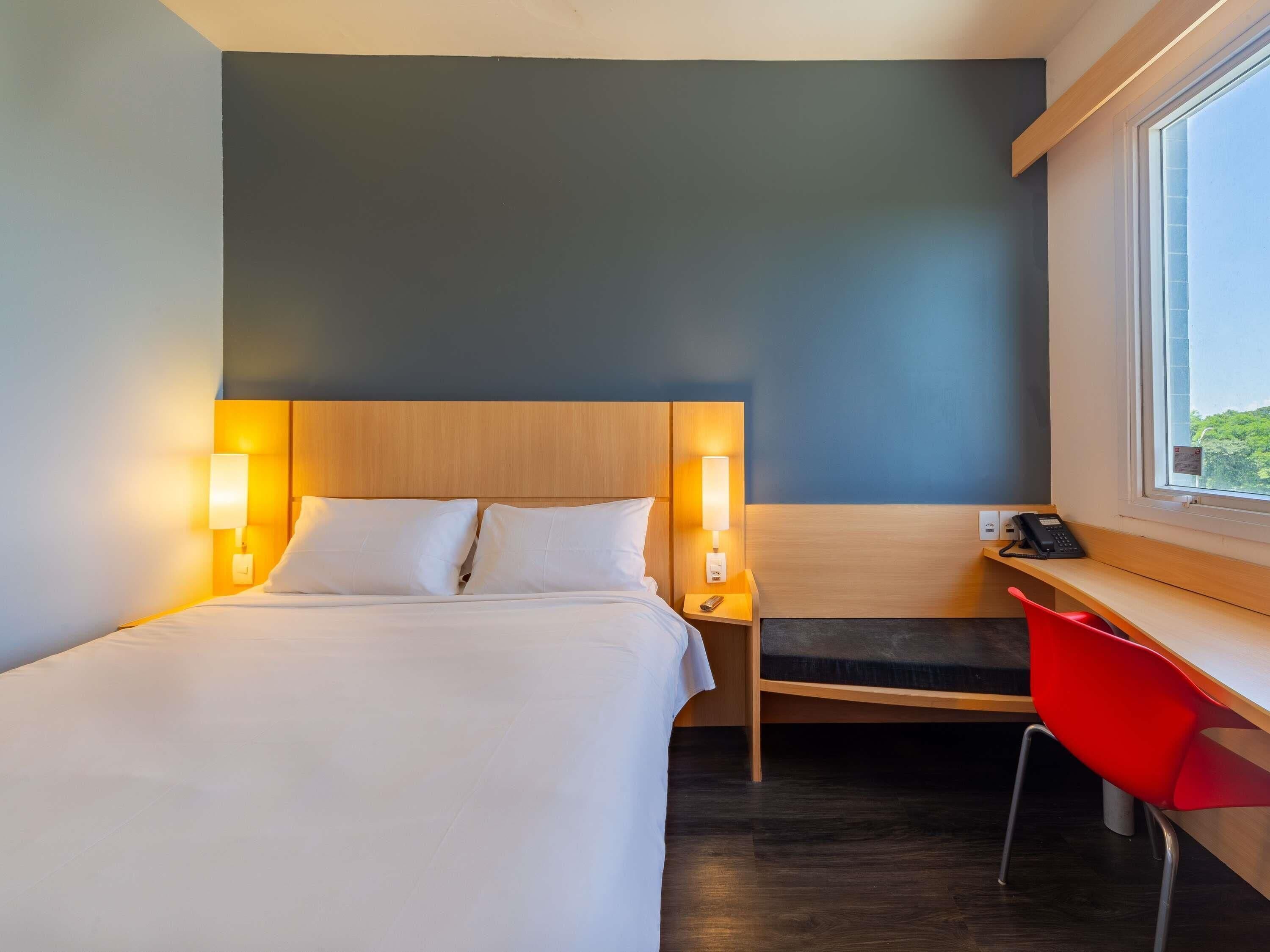 A hotel room with a bed, bedside lamps, a desk with a chair, and a window.