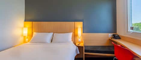Et hotelværelse med en seng, sengeborde med lamper, et skrivebord med stol og et vindue.