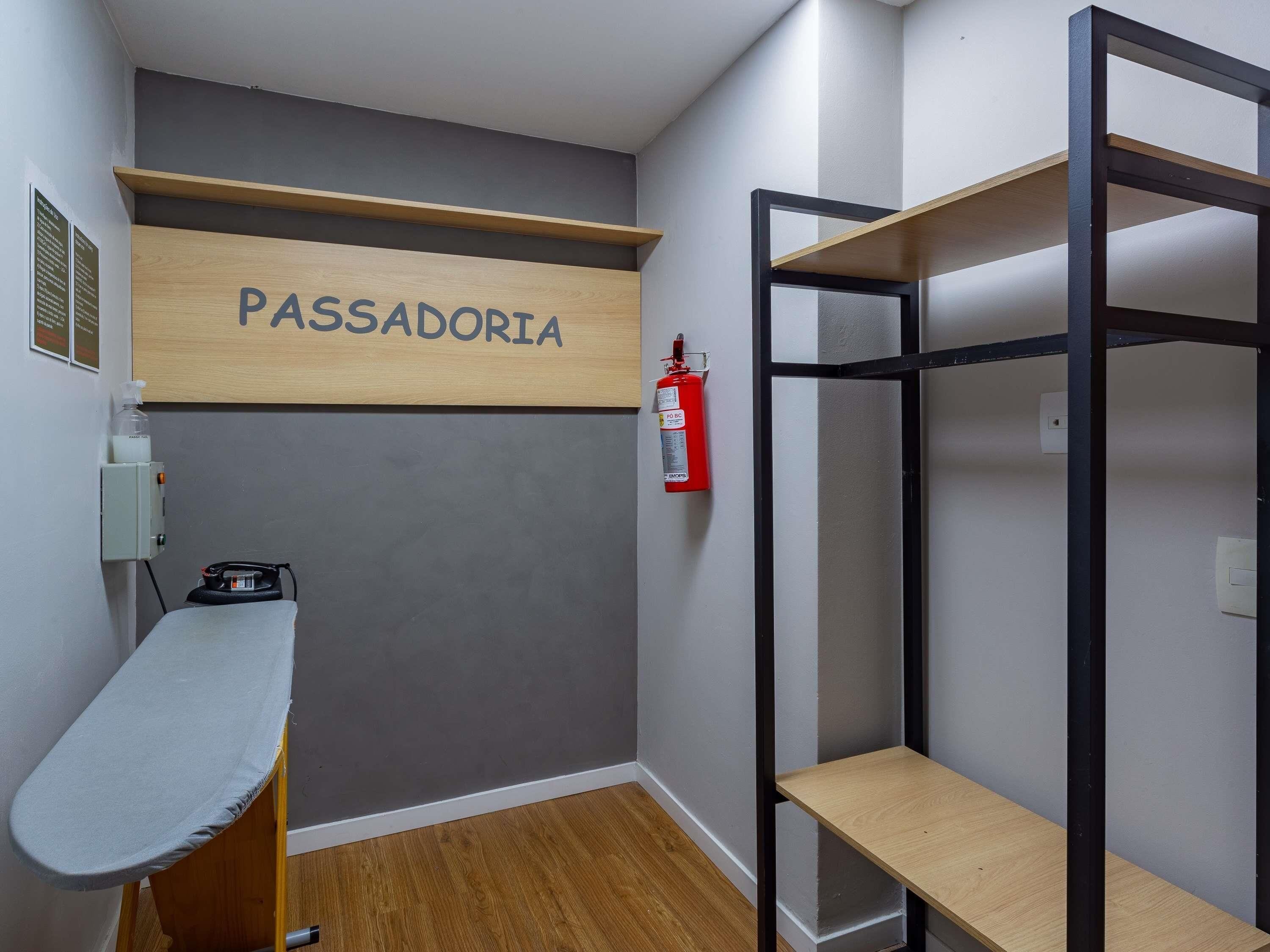 A room with a wooden shelf labeled 'PASSADORIA,' a fire extinguisher, an ironing board, and a bunk bed with a wooden shelf.