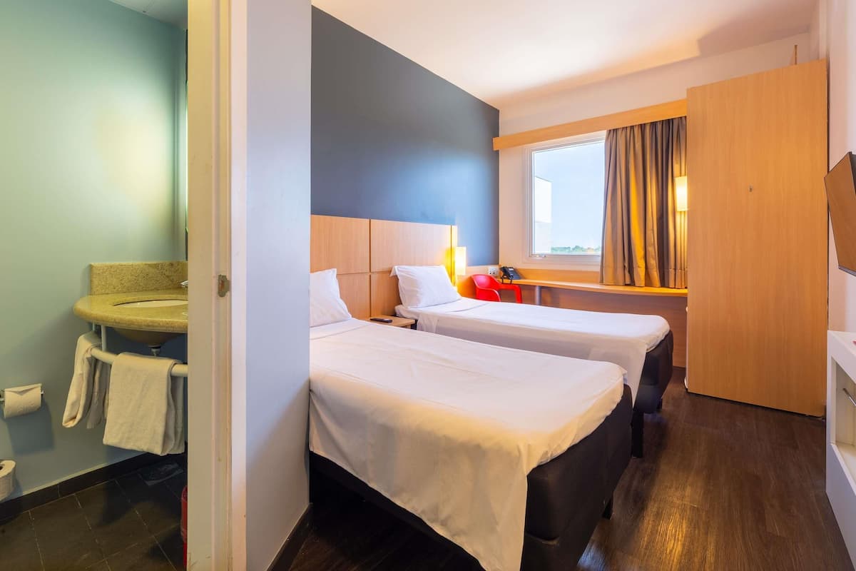 Une chambre d’hôtel avec deux lits, un bureau, une fenêtre avec des rideaux et une salle de bain équipée d’un lavabo et d’un porte-serviettes.