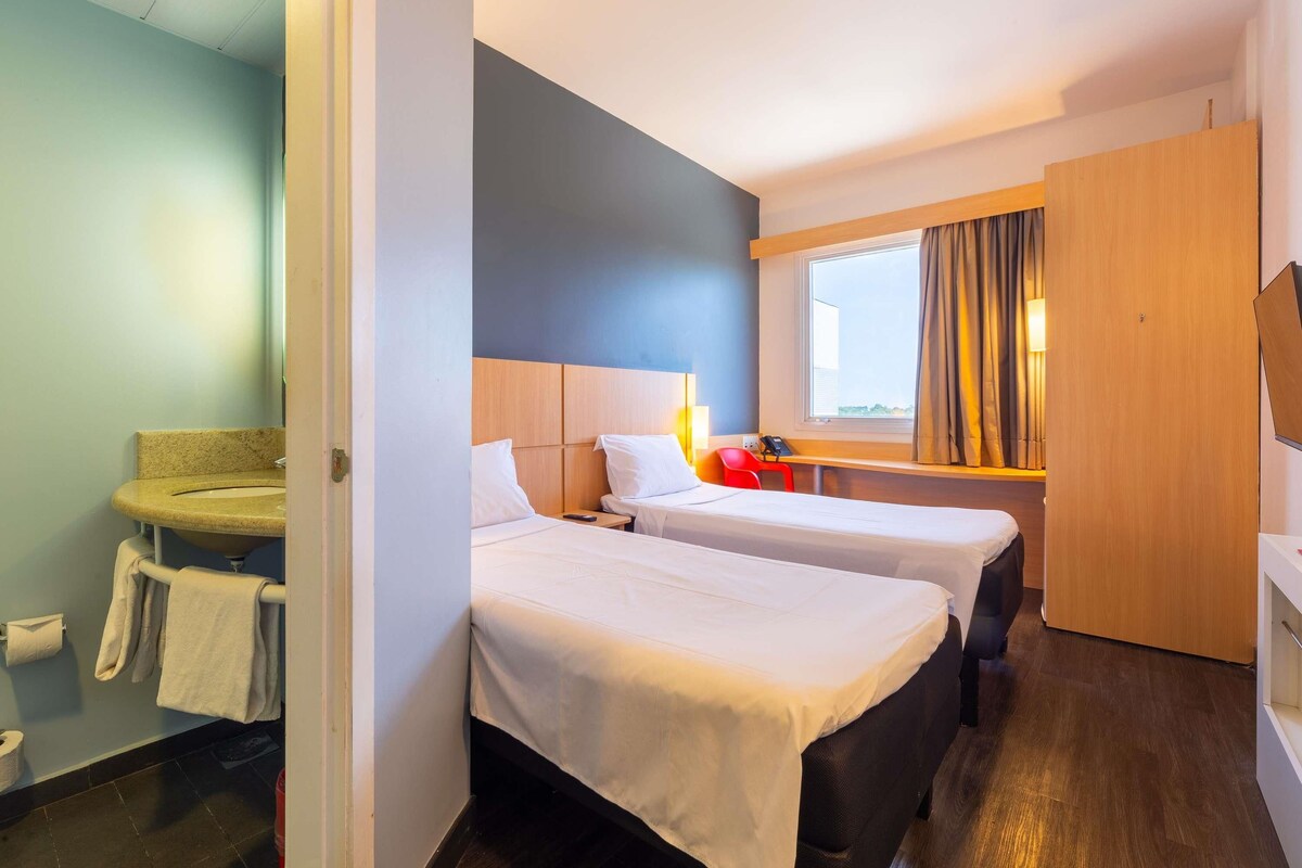 Habitación de hotel con dos camas, un escritorio, una ventana con cortinas y un baño con lavabo y toallero.