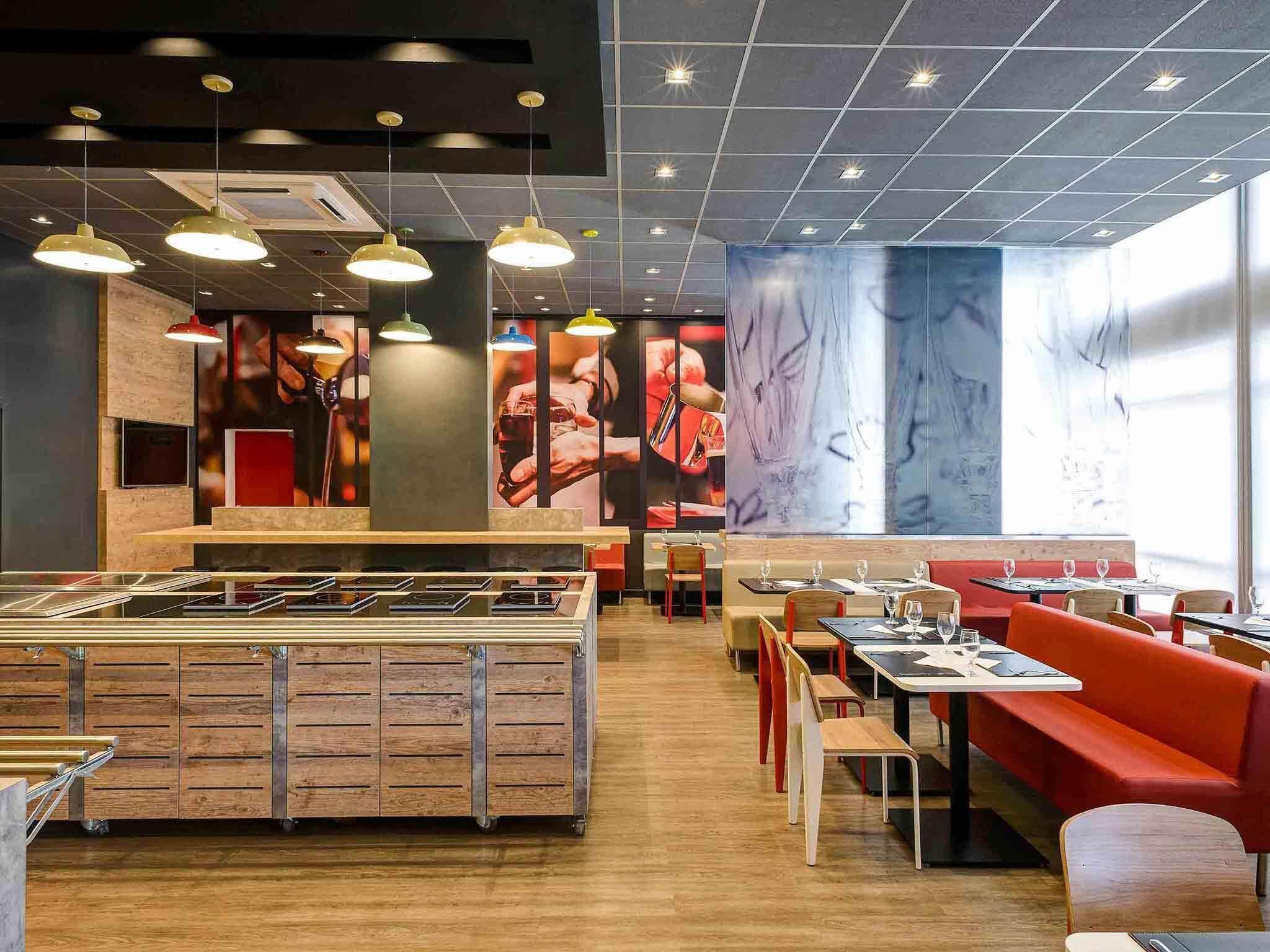 En restaurantindretning med en central ø, borde med siddepladser og hængende lamper.