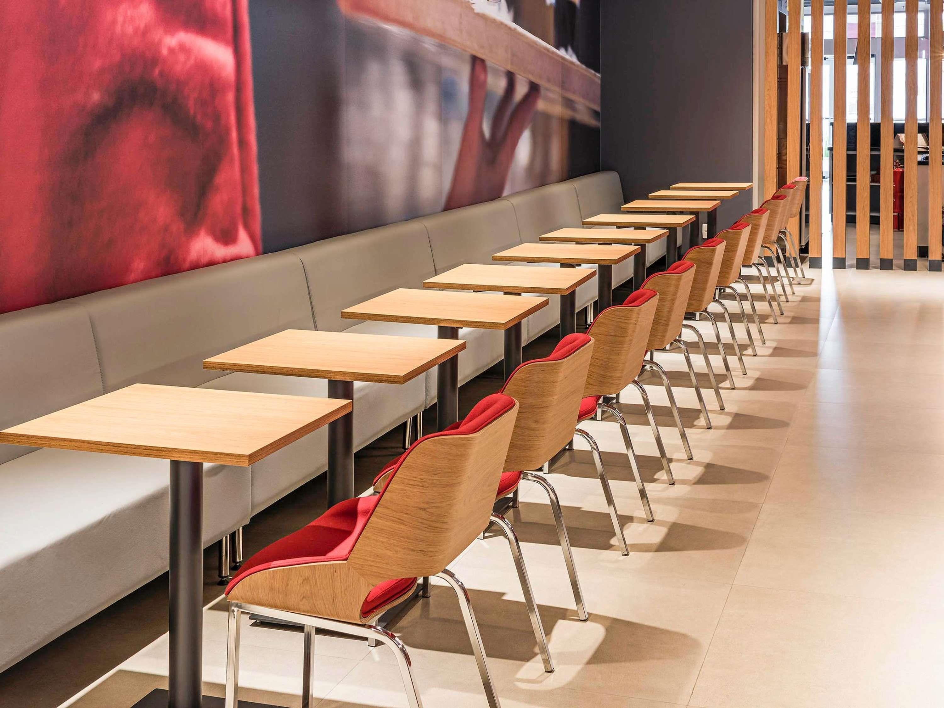 A modern dining area with long tables and red upholstered chairs, a grey bench, and a large abstract painting on the wall.