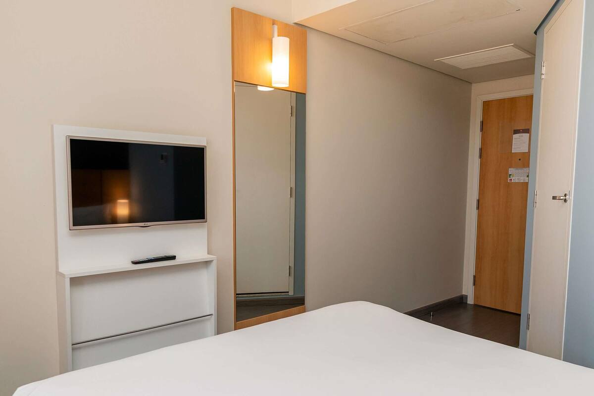 Una habitación de hotel con una cama, un televisor montado en la pared, un espejo y una puerta.