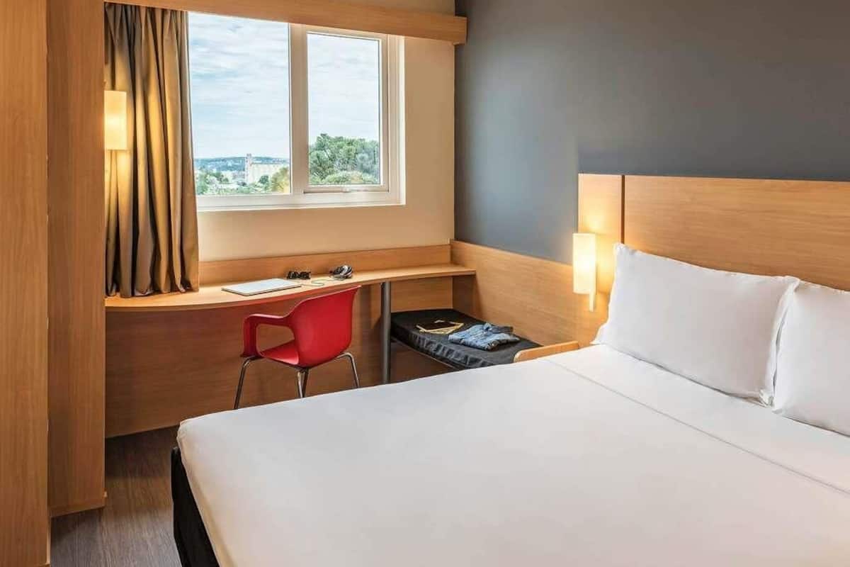 A hotel room with a bed, a desk, a chair, a window, and a lamp.