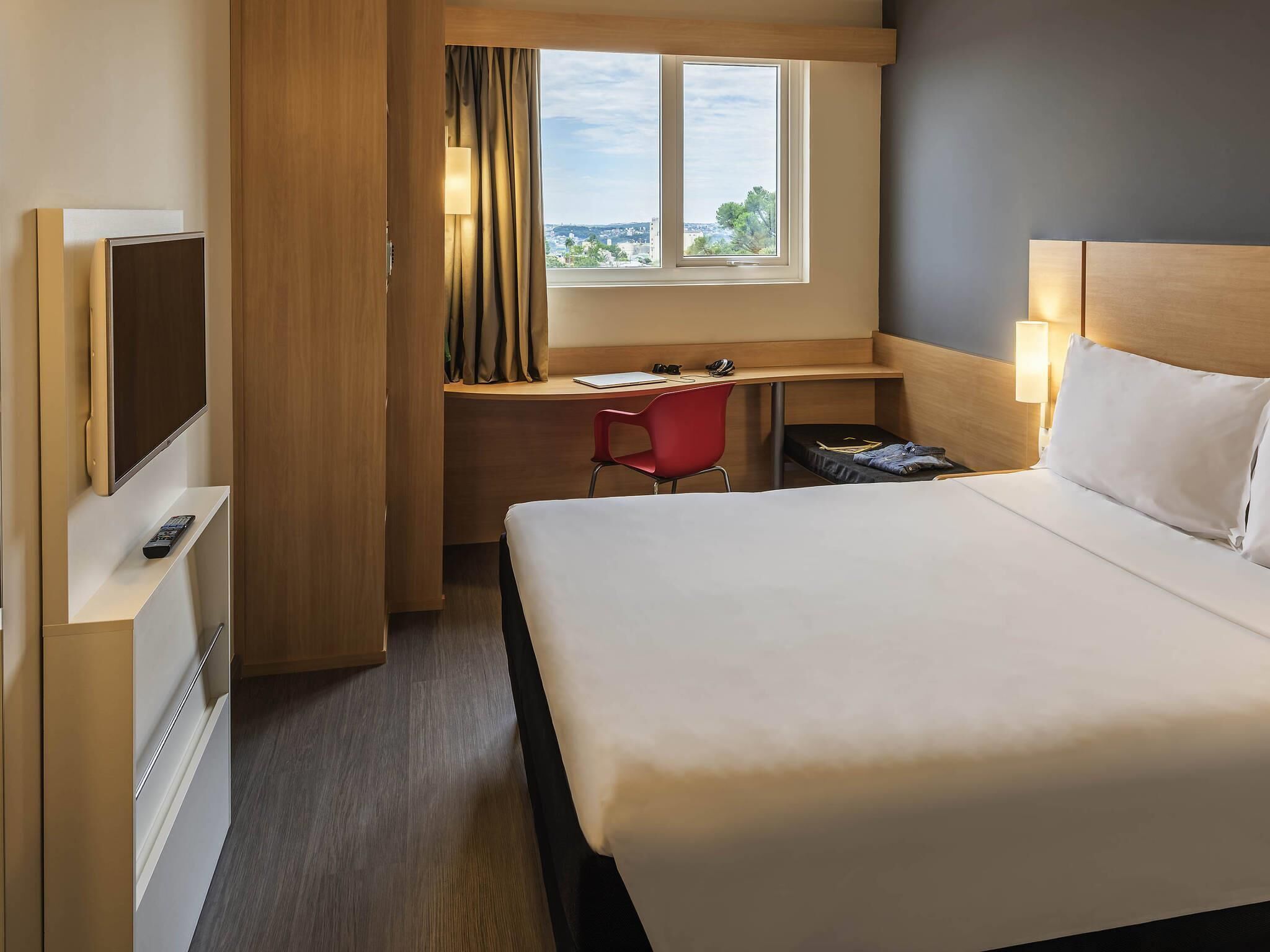 A hotel room with a bed, a desk with a chair, a TV, and a window with curtains.