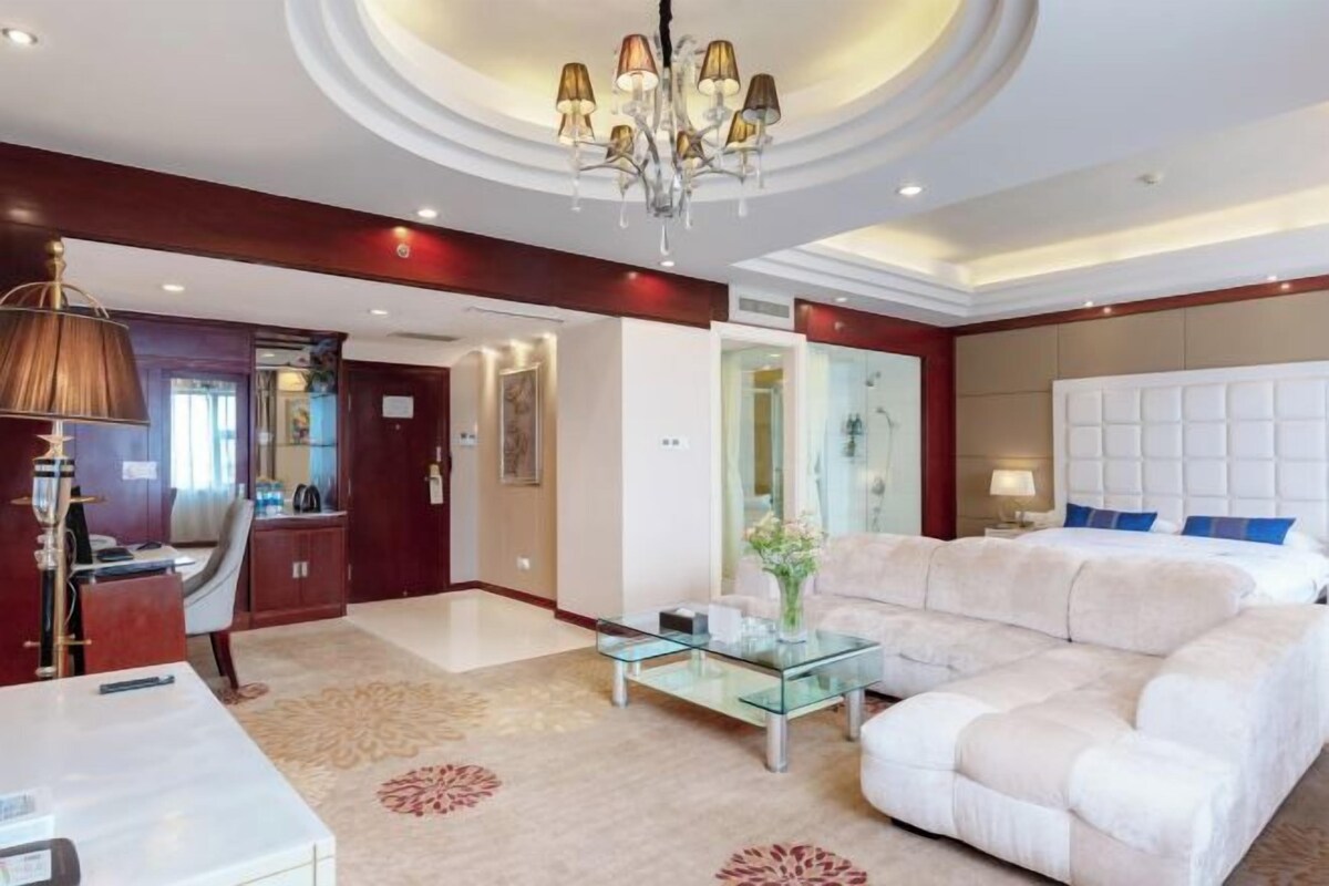 A hotel room with a large bed, a sofa, a glass coffee table, a desk with a lamp, and a chandelier.