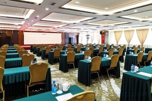 Un salón de conferencias con mesas y sillas dispuestas en filas, una pantalla de proyección y suelo alfombrado.