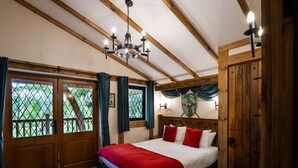 A bedroom with a wooden ceiling, a chandelier, a bed with red and white bedding, and a view through a window with curtains. -  Warwick Castle Hotel and Knight's Village (Warwick)