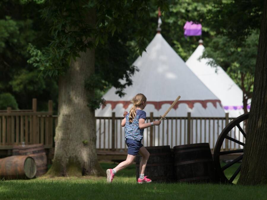 A child playing with a toy sword in a park with a white tent and wooden barrels in the background.