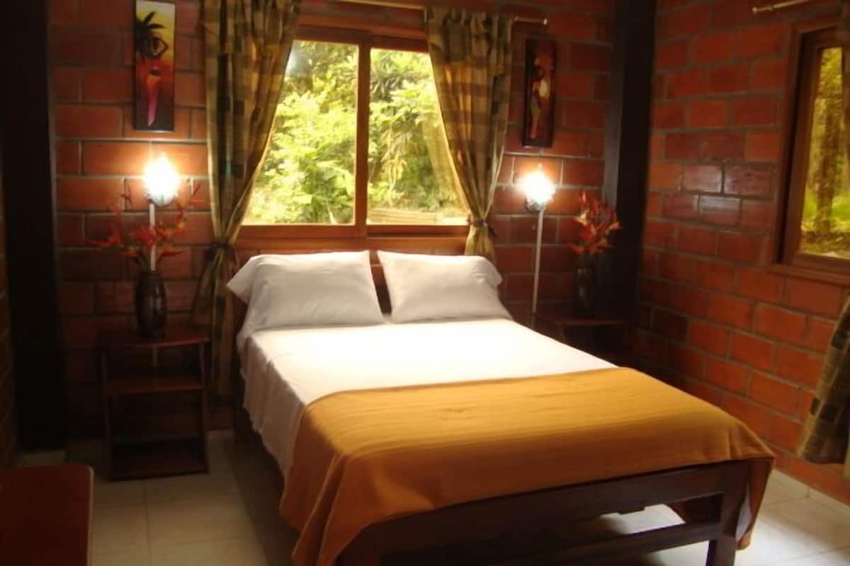 Un lit simple dans une pièce aux murs en briques, une fenêtre avec des rideaux et deux lampes fixées au mur.