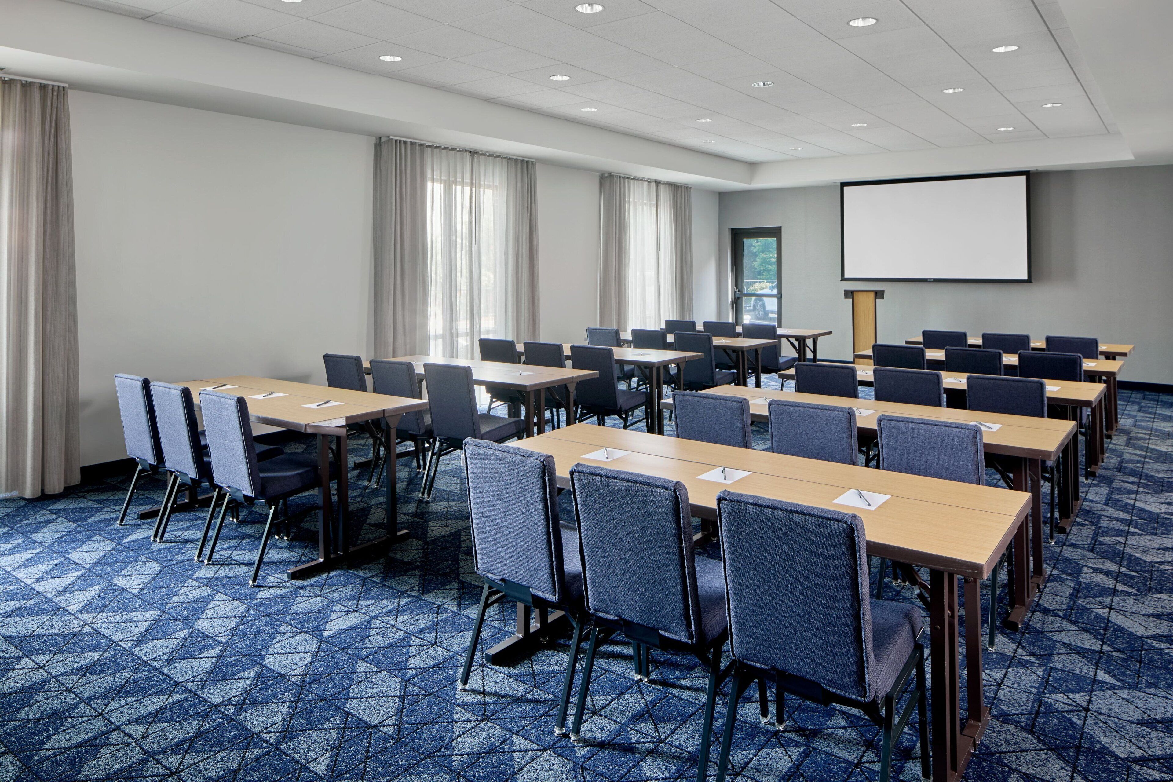 A conference room with multiple tables and chairs, a projector screen, and a large window with curtains.
