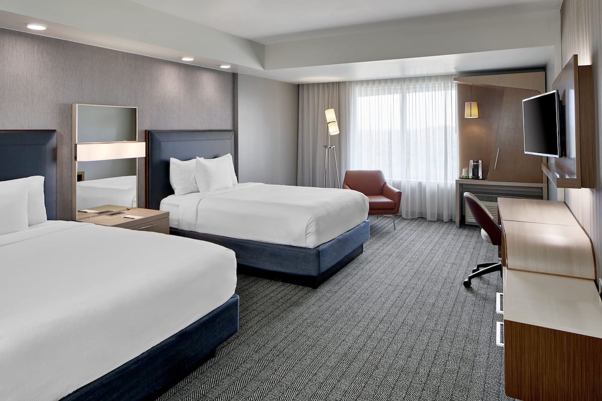 A hotel room with two beds, a desk, a chair, a TV, and a large window with curtains.