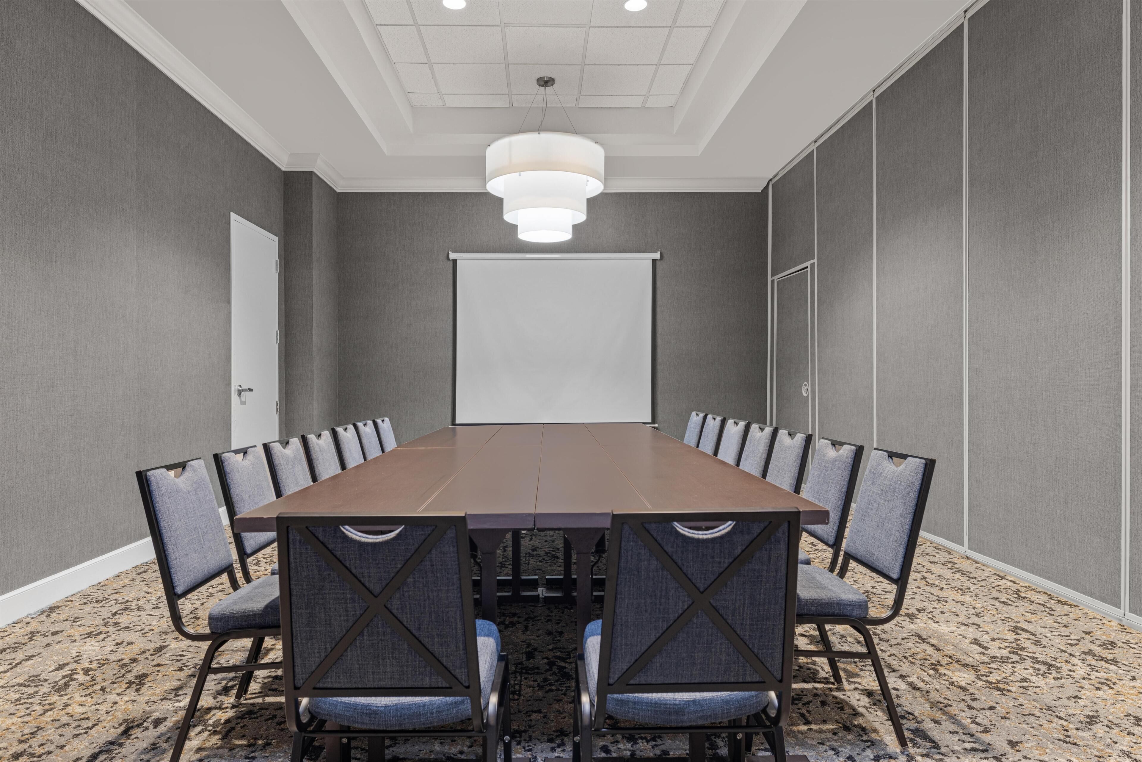 Een vergaderzaal met een lange tafel, stoelen, een projectiescherm en een plafondlamp.