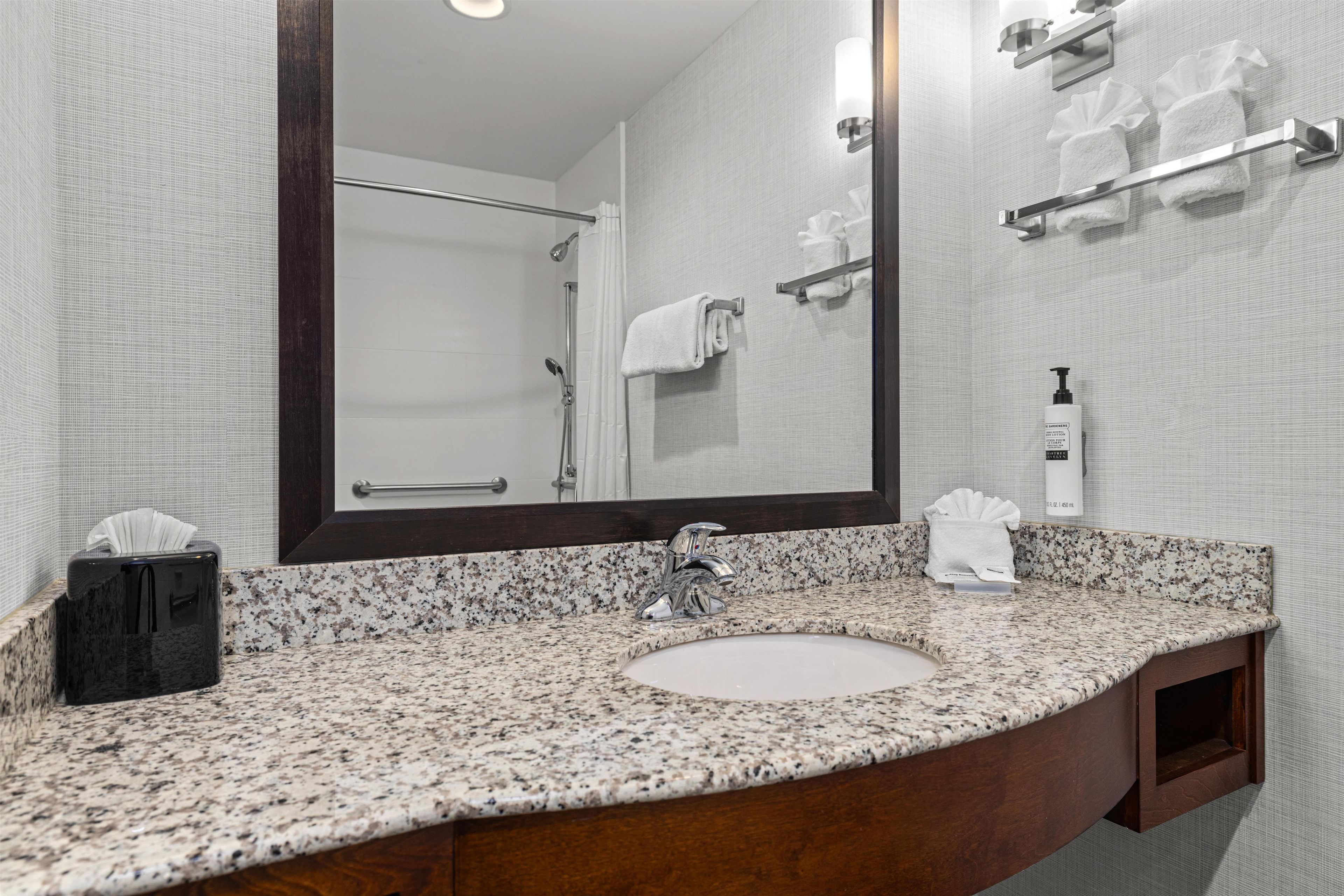 Une salle de bain avec un lavabo doté d’un plan de travail en granit, un miroir, un porte-serviettes et une bouteille de shampoing.