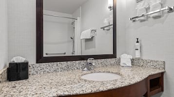 Une salle de bain avec un lavabo doté d’un plan de travail en granit, un miroir, un porte-serviettes et une bouteille de shampoing.