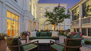 Un espacio exterior con mobiliario de jardín, incluyendo un sofá y sillas verdes, rodeado de plantas en macetas y un árbol.