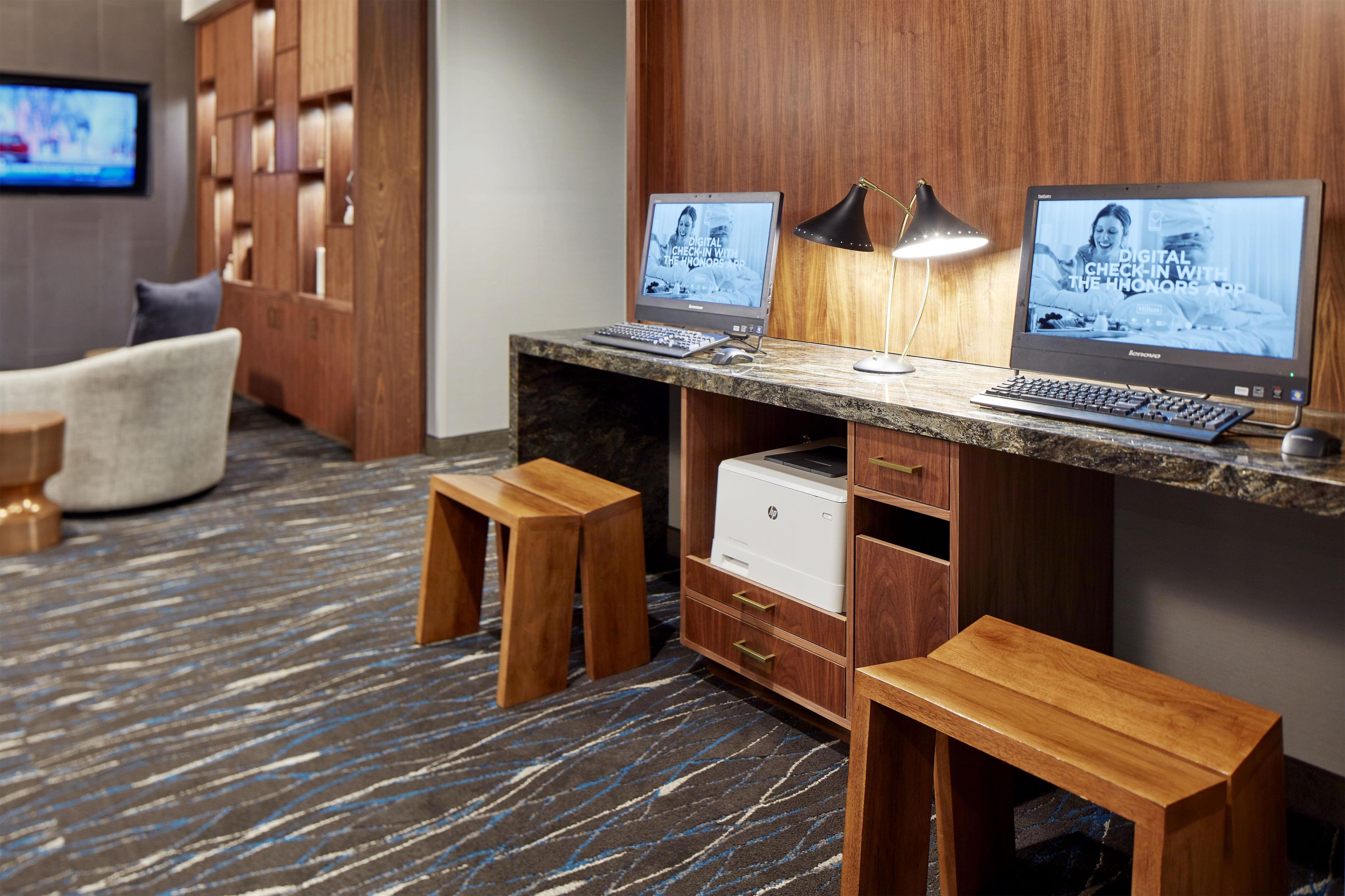A hotel workspace with two desktop computers, a printer, and a lamp on a wooden desk.