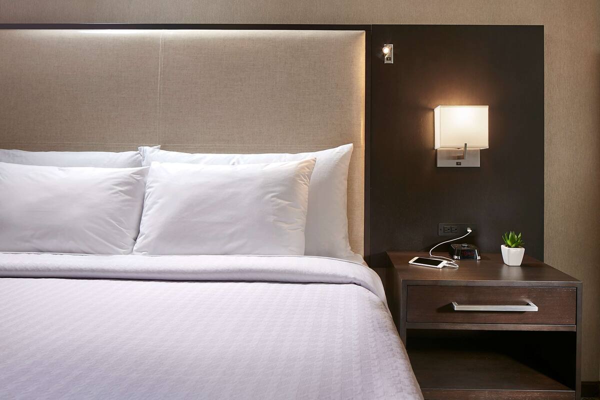 A neatly made bed with white linens, a bedside table with a lamp, a small potted plant, and a smartphone charging on the table.