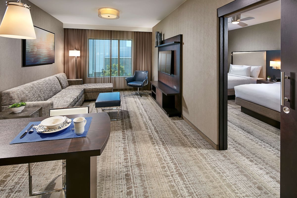 A hotel room with a sofa, a TV, a bed, a dining table with a plate and a cup, and a window with curtains.