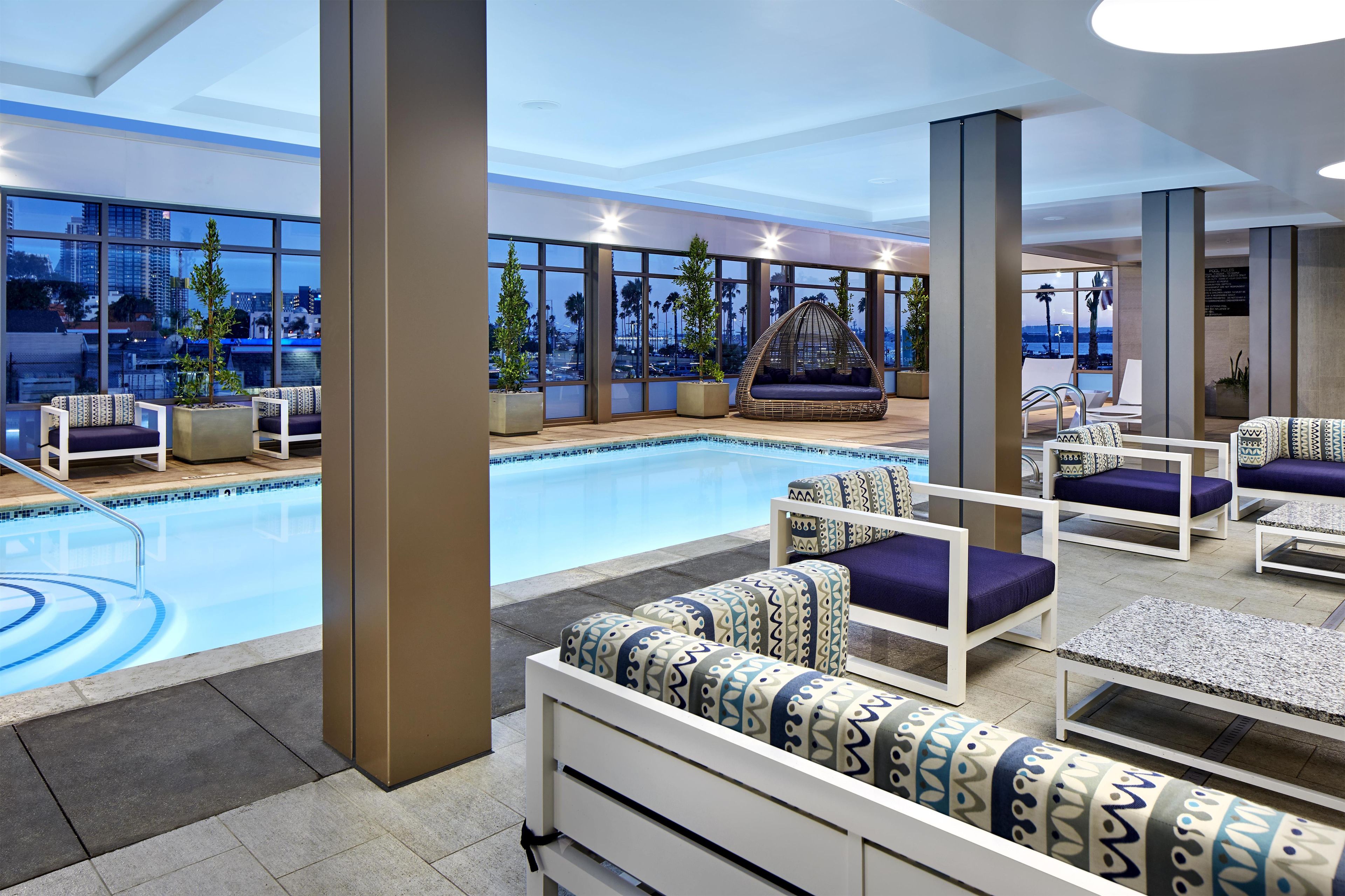 Uma área interna de piscina moderna, com assentos e amplas janelas que oferecem vista para a paisagem urbana.